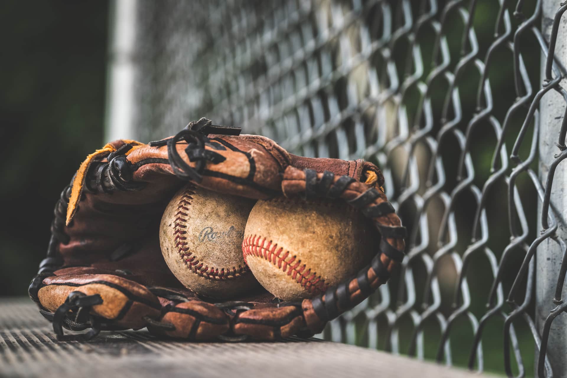 A worn leather fielding glove holding two baseballs beside a chain-link fence